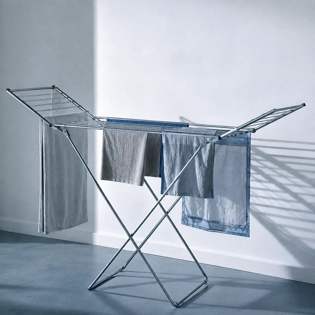 A folding indoor airer against a white wall, thin rails casting long parallel shadows.