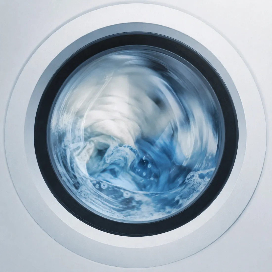 The round porthole of a washing machine mid-cycle, pale textile blurred behind the glass.